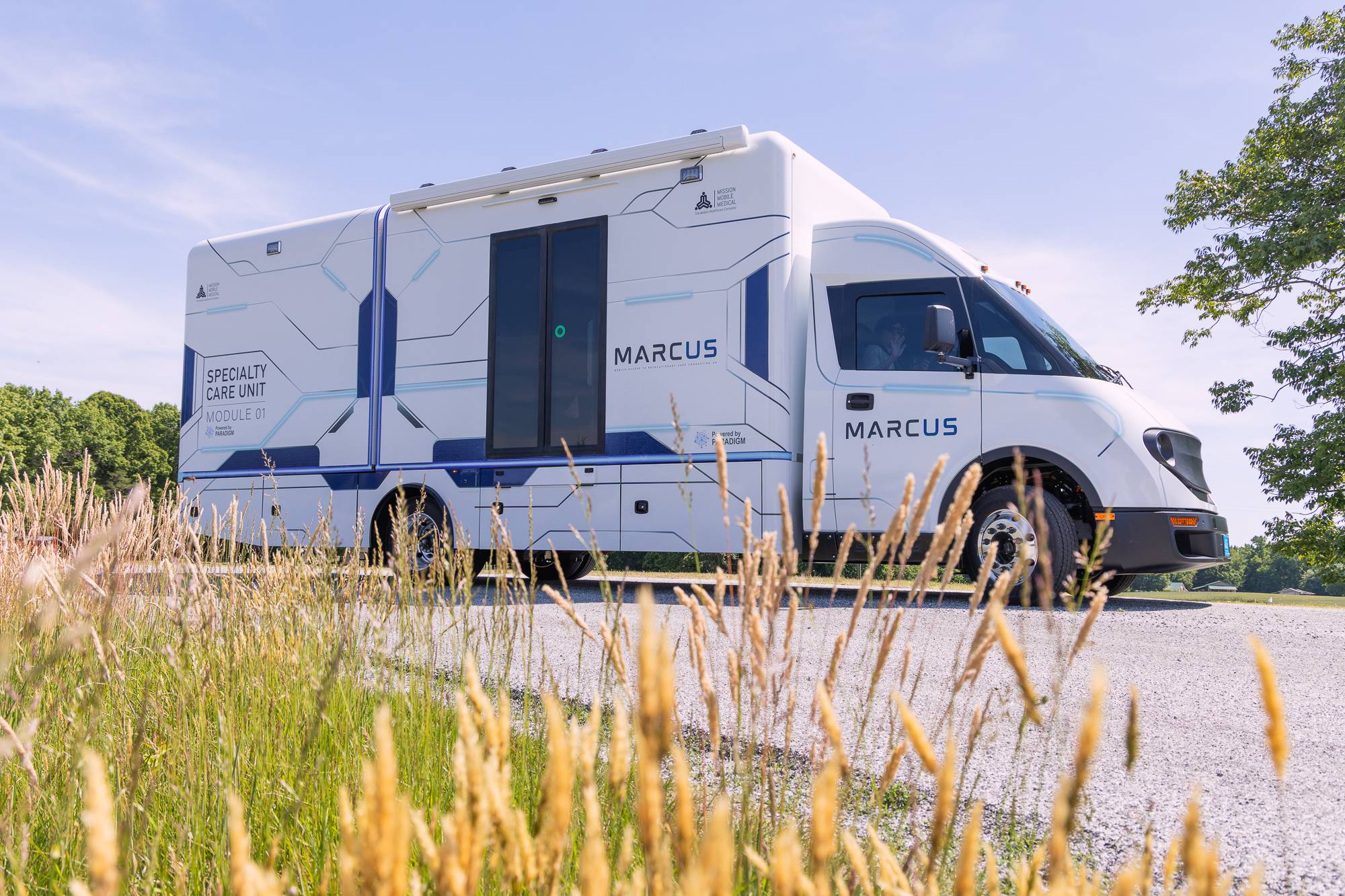 MARCUS parked outdoors on a gravel lot with summer grass in the foreground